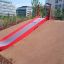 Rote Hangrutsche Breit SLW180 mit Metallrutsche auf Spielplatz in urbanem Umfeld.