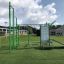 Spielplatzgerät New Age Fit grün mit Kletterstangen, Ringen und Trainingsstation auf Kunstrasen.
